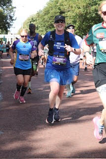Amanda, a PFM Coaching athlete, running mid-race at the 2026 Cardiff Half Marathon in a blue top, black cap and running vest, alongside other runners on a tree-lined road